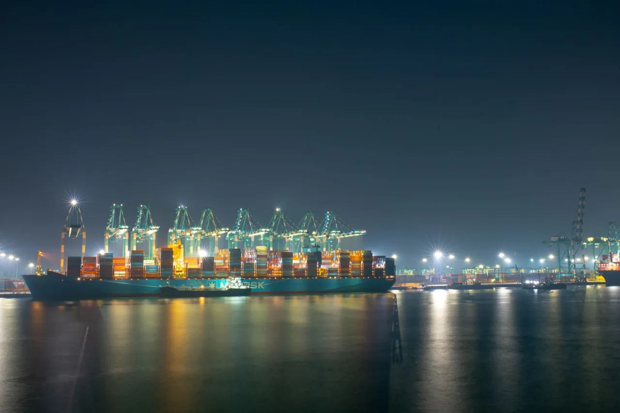 A cargo ship docked at a bustling port, illuminated at night with cranes and containers.