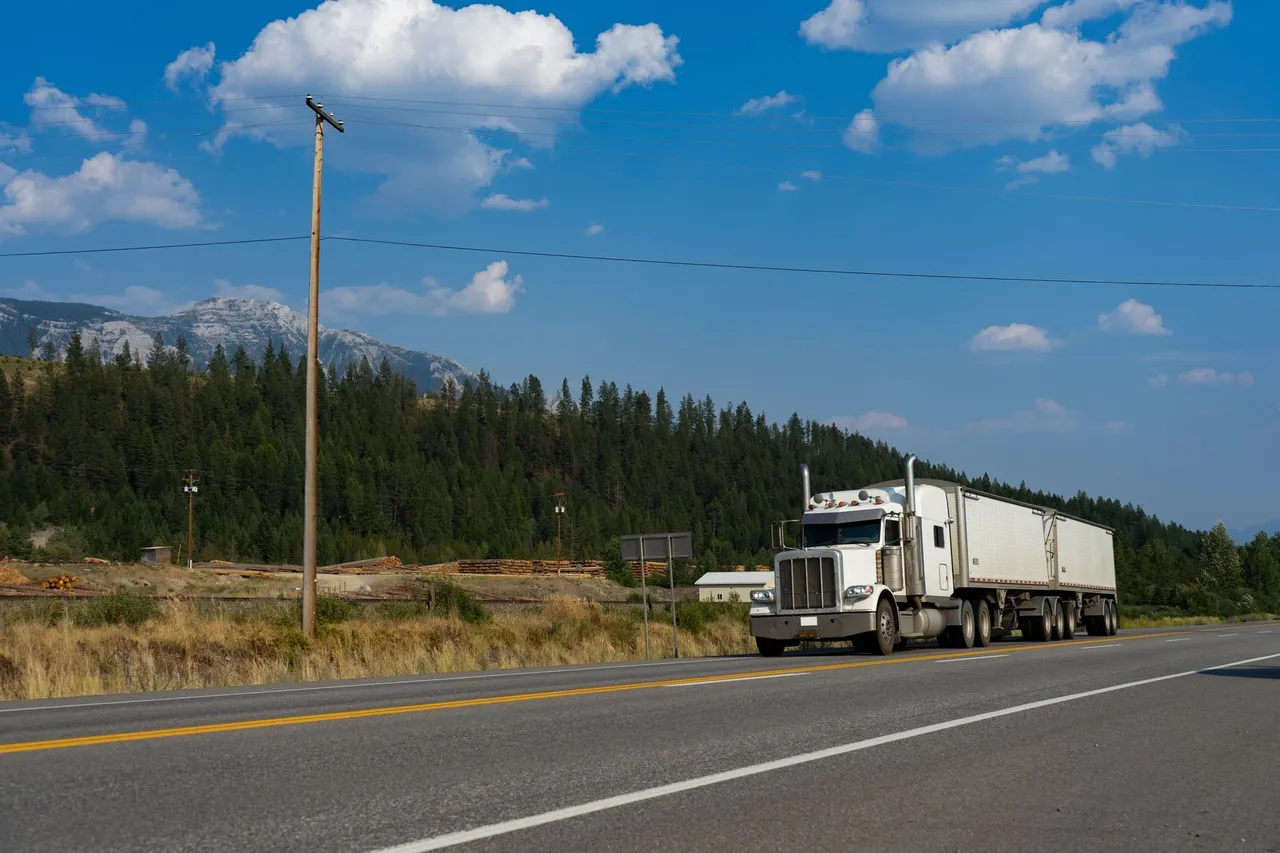 A semi-truck travels on a scenic highway with mountains in the background under a clear sky.