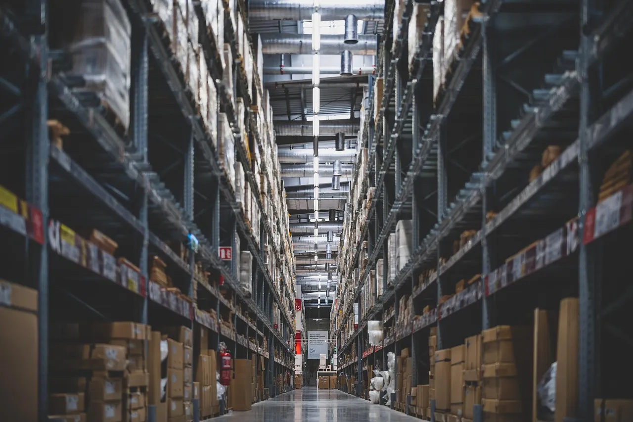 Two workers handle a package in a spacious warehouse surrounded by shelves stocked with boxes and products.